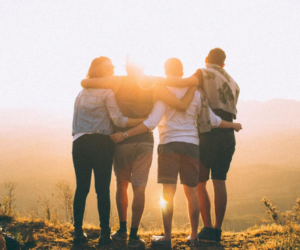 four people with their arms around each other's shoulders watching a sunrise from a mountaintop