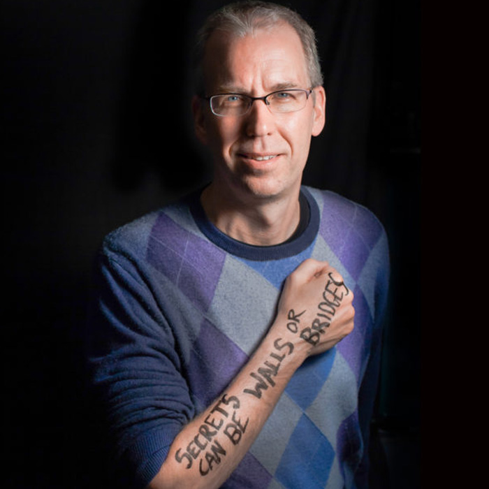 A man with short gray hair and glasses stands in front of a black background wearing an argyle sweater. He holds his hand in a fist across his chest, with the words