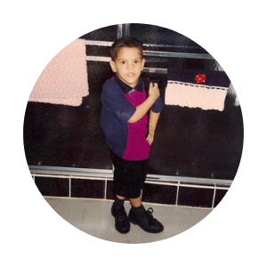 A baby photo of Mark, in a bathroom, posing with his arm on his hip and his other arm on his shoulder with his legs crossed and inverted. He has short black hair and is wearing black shorts, a navy and pink shirt, and black shoes.