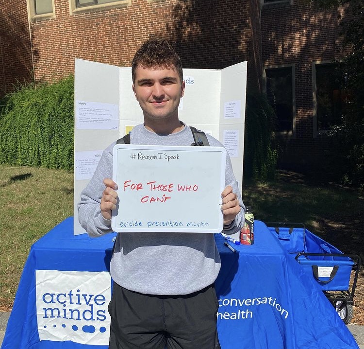 A young adult in front of an Active Minds table holding a dry erase board that reads I speak for those who cant, suicide prevention month